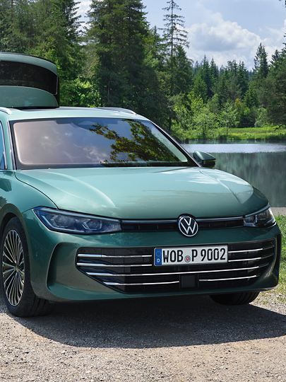 VW Der neue Passat VW Passat in blaugrün parkt an einem Badesee mit geöffnetem Kofferraum. Ein Mann verteilt Freitzeitspielzeug an zwei Kinder.